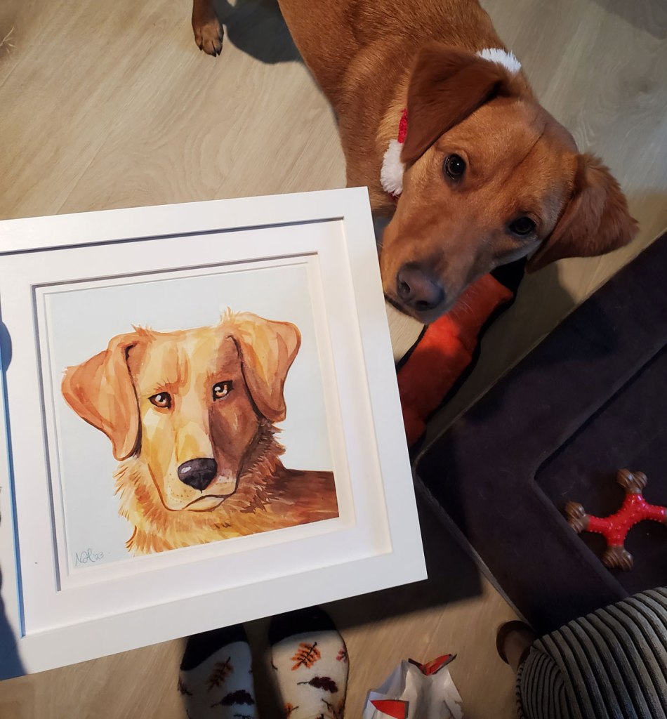 A photo of the frame watercolour dog portrait, being held next to the dog himself. He is sniffing the frame, looking curious. 