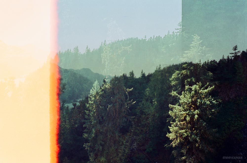A film multiple exposure photo with a light leak which makes part of the image look bright orange. The multiple exposure is of mountains and forests, the two layers of images blurring together. 
