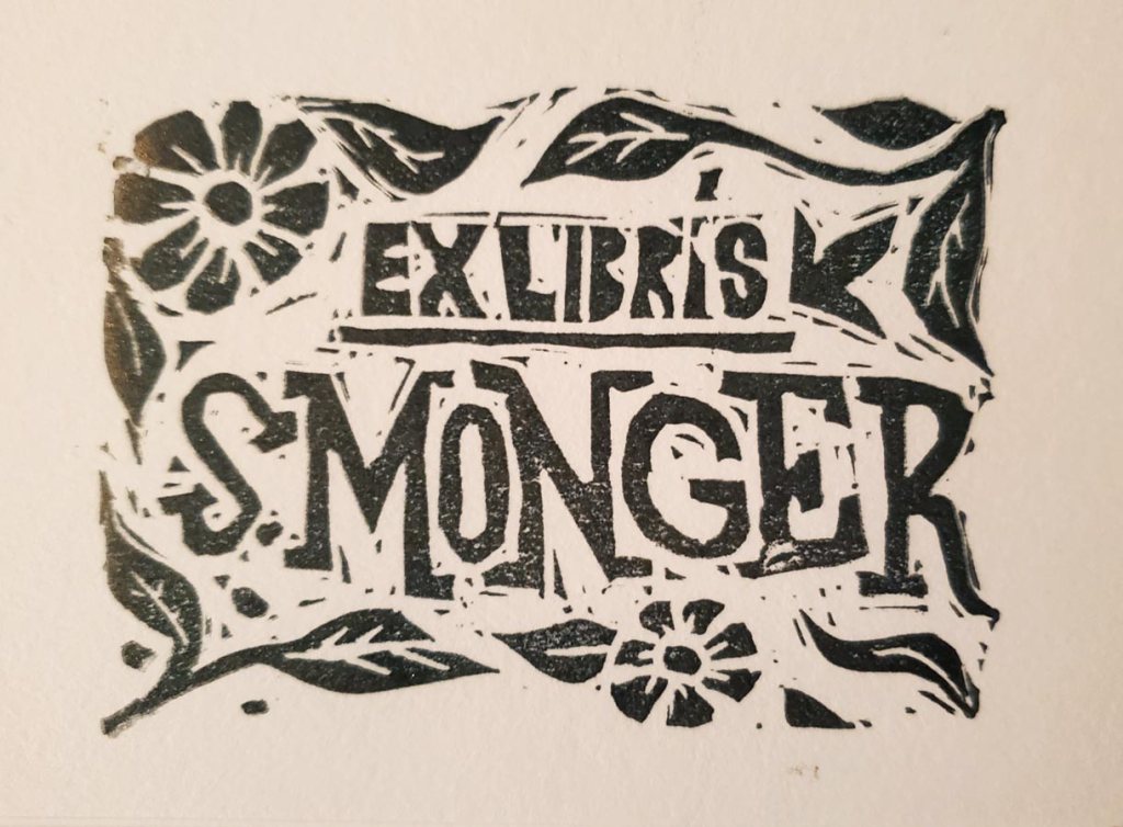 A photo of a Lino block print in black ink. The print reads: “Ex Libris S. Monger” – it’s a bookplate, and around the text are decorative vines and flowers.
