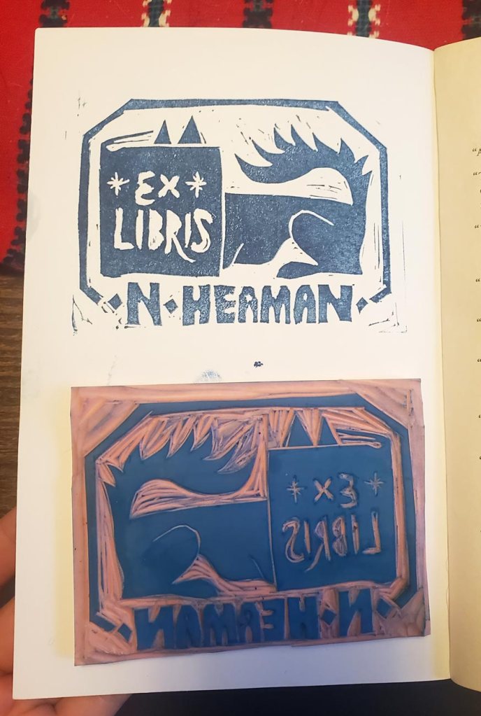 A photo of a bookplate Lino block siting next to a print of it on the inner cover of a book. This bookplate reads “Ex Libris N. Heaman” and has an illustration of a dog reading a book: the dog is laying down and her head is deep in an open book – only the tips of her pointy ears are seen poking up. 
